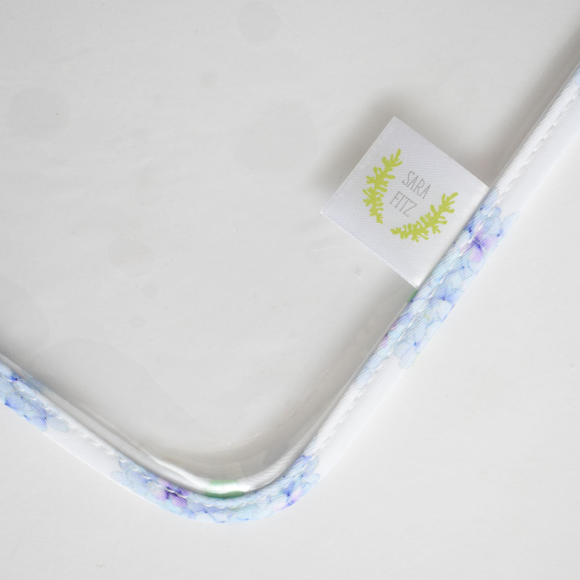 A close-up of a floral-patterned white frame with a Sara Fitz tag featuring green foliage, set against a white background—perfect for complementing Sara Fitz prints or pairing with your favorite Hydrangea Project Bag by Sara Fitz, crafted from heavyweight canvas.