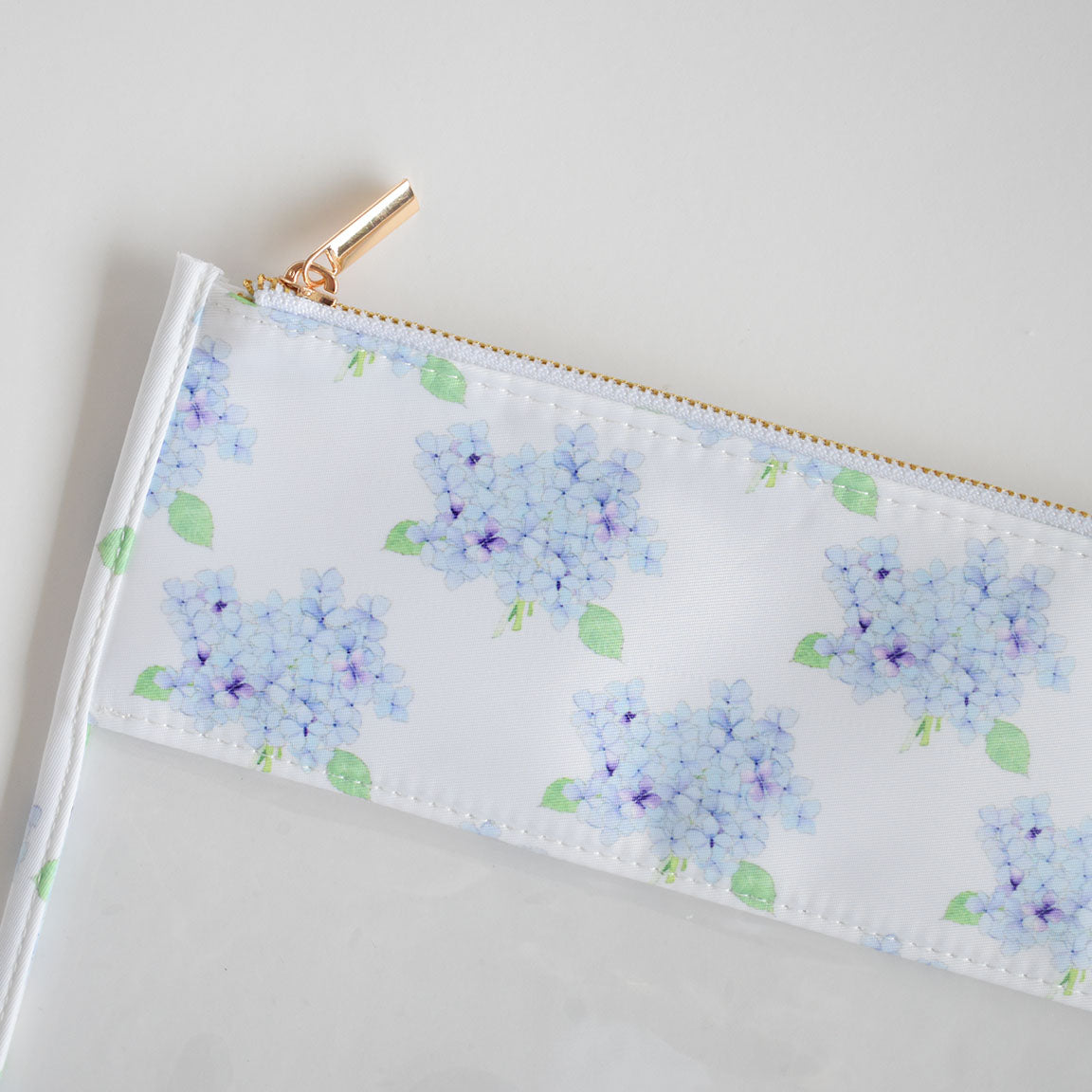 The Hydrangea Project Bag by Sara Fitz is a clear pouch with a white top section adorned with signature Sara Fitz prints of light blue hydrangea flowers and green leaves, crafted from heavyweight canvas and completed with a gold zipper.
