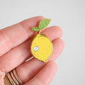 A hand holds the Sara Fitz Lemon Needle Minder, a small enamel pin shaped like a yellow lemon with a leaf—ideal as an enamel needle minder or stitching accessory. The lemon features a tiny sticker that says "fresh," and the person is wearing several rings on their fingers.