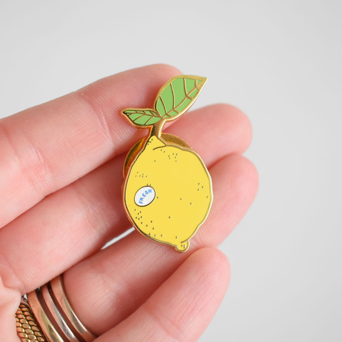 A hand holds the Sara Fitz Lemon Needle Minder, a small enamel pin shaped like a yellow lemon with a leaf—ideal as an enamel needle minder or stitching accessory. The lemon features a tiny sticker that says "fresh," and the person is wearing several rings on their fingers.