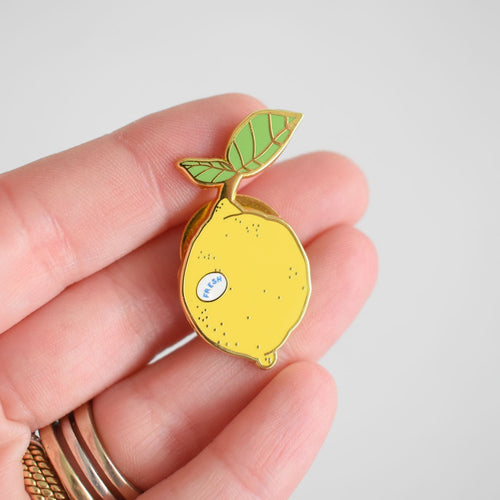 A hand holds the Sara Fitz Lemon Needle Minder, a small enamel pin shaped like a yellow lemon with a leaf—ideal as an enamel needle minder or stitching accessory. The lemon features a tiny sticker that says "fresh," and the person is wearing several rings on their fingers.