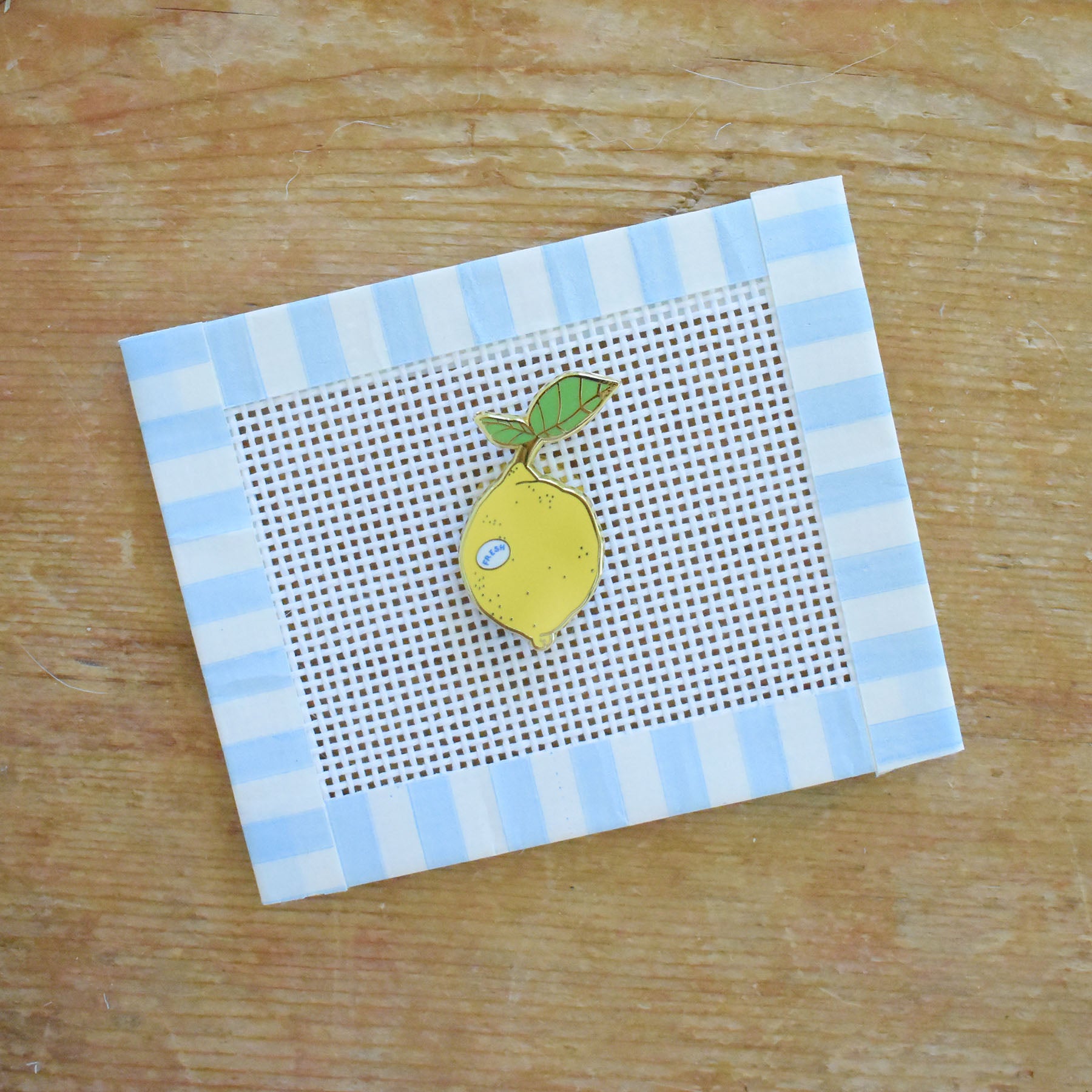 A small needlepoint canvas with a blue and white striped border features a stitched yellow lemon with a green leaf, placed on a wooden surface. The Sara Fitz Lemon Needle Minder pairs beautifully with this charming piece, making it a delightful stitching accessory for any crafter.