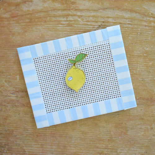 A small needlepoint canvas with a blue and white striped border features a stitched yellow lemon with a green leaf, placed on a wooden surface. The Sara Fitz Lemon Needle Minder pairs beautifully with this charming piece, making it a delightful stitching accessory for any crafter.