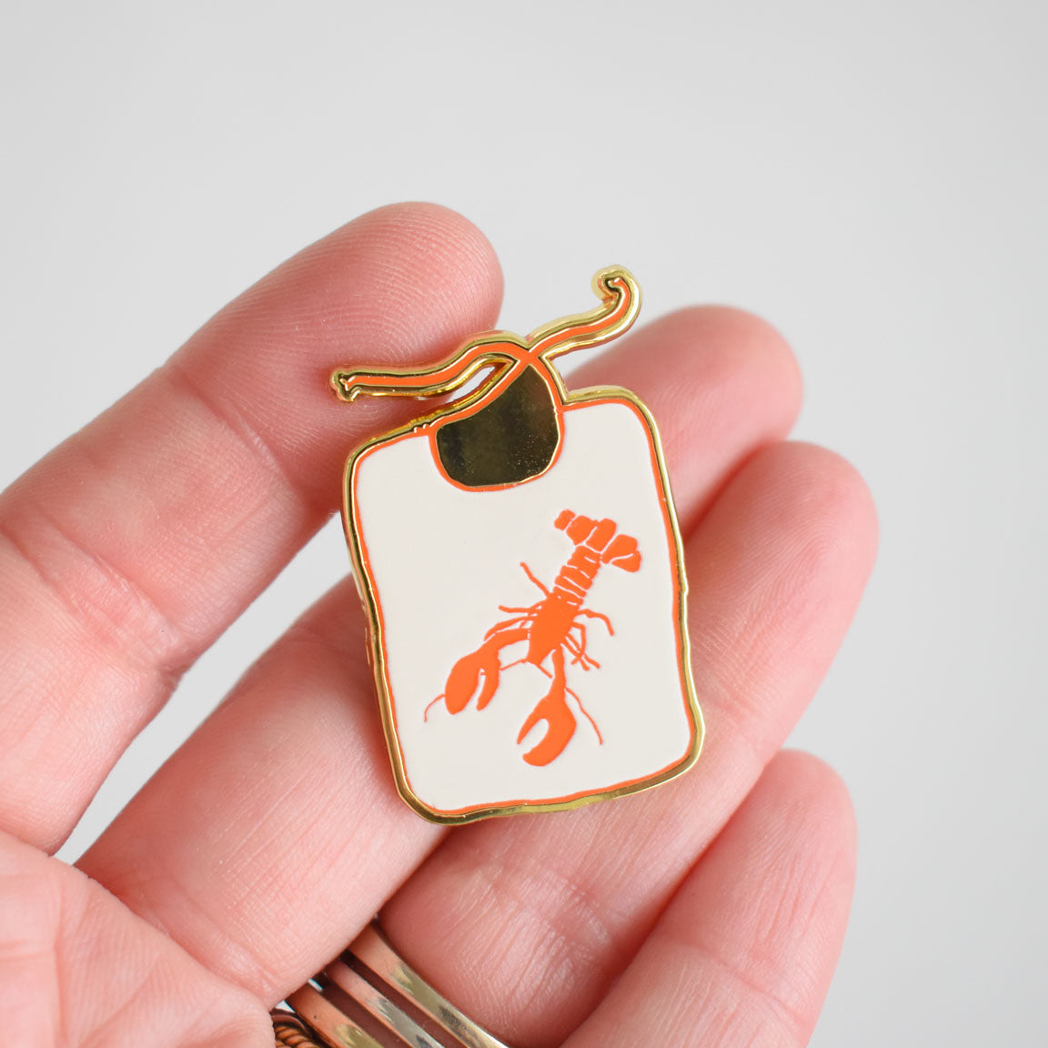 A hand holds the Sara Fitz Lobster Bib Needle Minder, a small enamel pin shaped like a white cutting board with a gold outline and an orange lobster illustration at its center, making it a charming and unique stitching accessory.