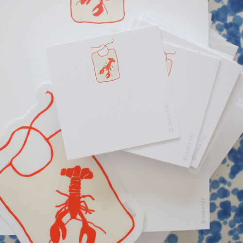 The Lobster Bib Notelet by Sara Fitz features a stack of square white cards with a red lobster bib illustration, plus a larger matching sticker, all displayed on a white surface with blue speckles. Printed in the USA.