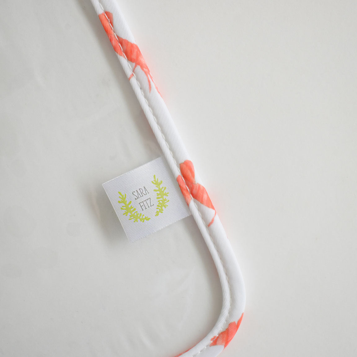A white fabric Lobster Project Bag by Sara Fitz, featuring a pink flamingo print and a small white tag with "Sara Fitz" in green text, lying on a plain white surface.