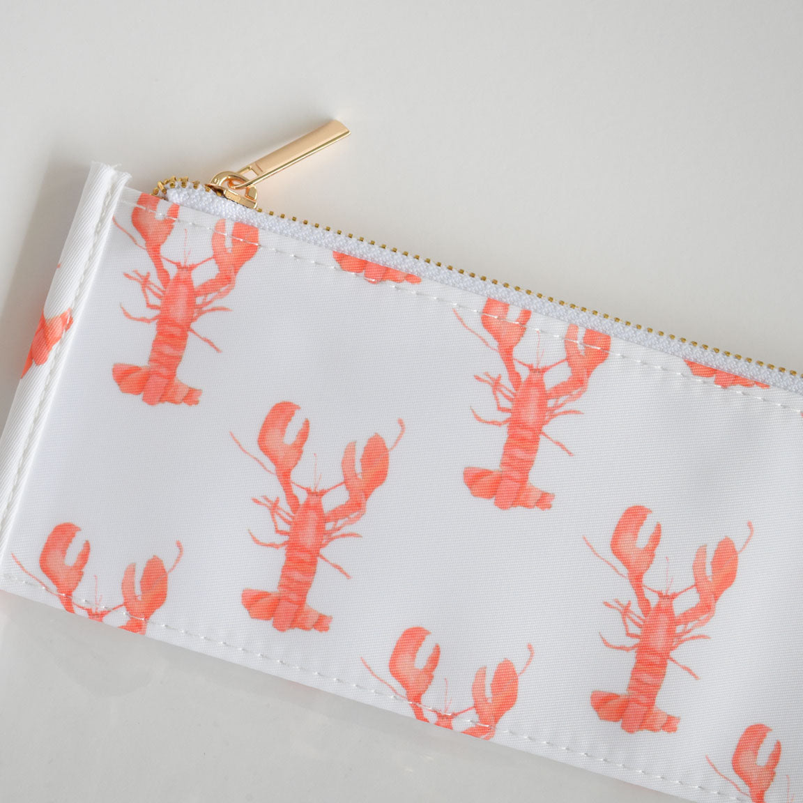 The Lobster Project Bag by Sara Fitz is a white zippered pouch featuring a gold zipper and an all-over red lobster pattern.