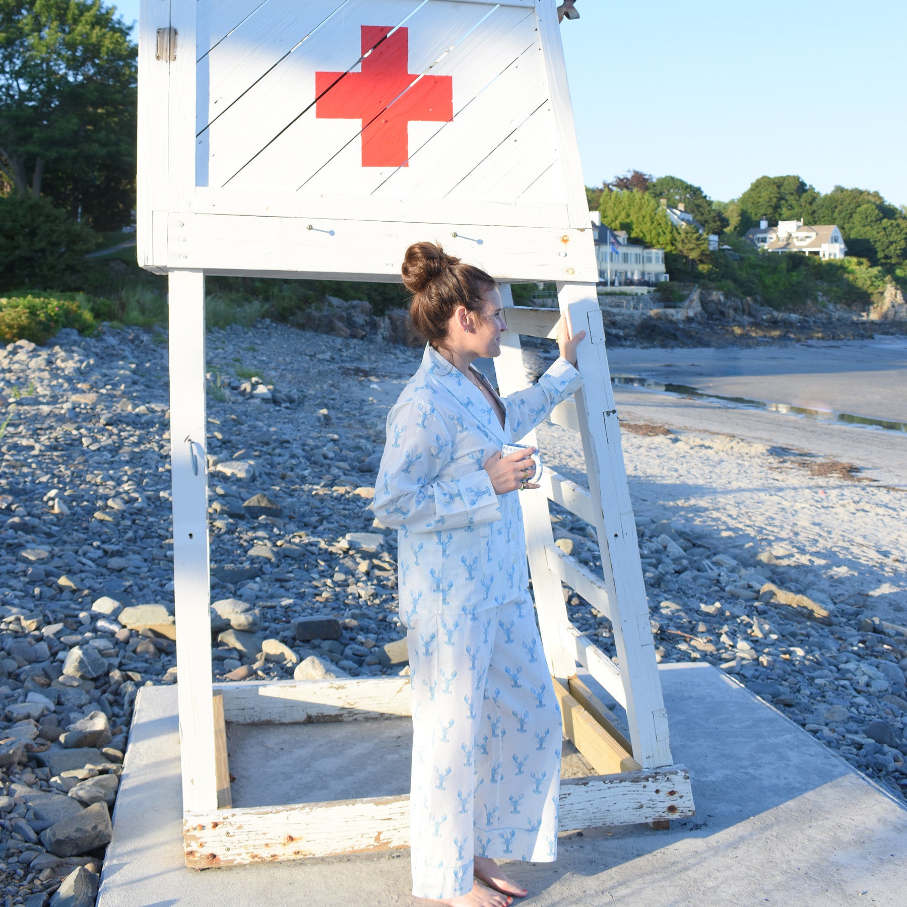 Wearing the Blue Lobster Pants Pajama Set by Sara Fitz, a woman stands barefoot by a white wooden lifeguard stand with a red cross, holding a mug on a rocky beach lined with houses and trees—perfectly capturing an effortless coastal sleepwear style.