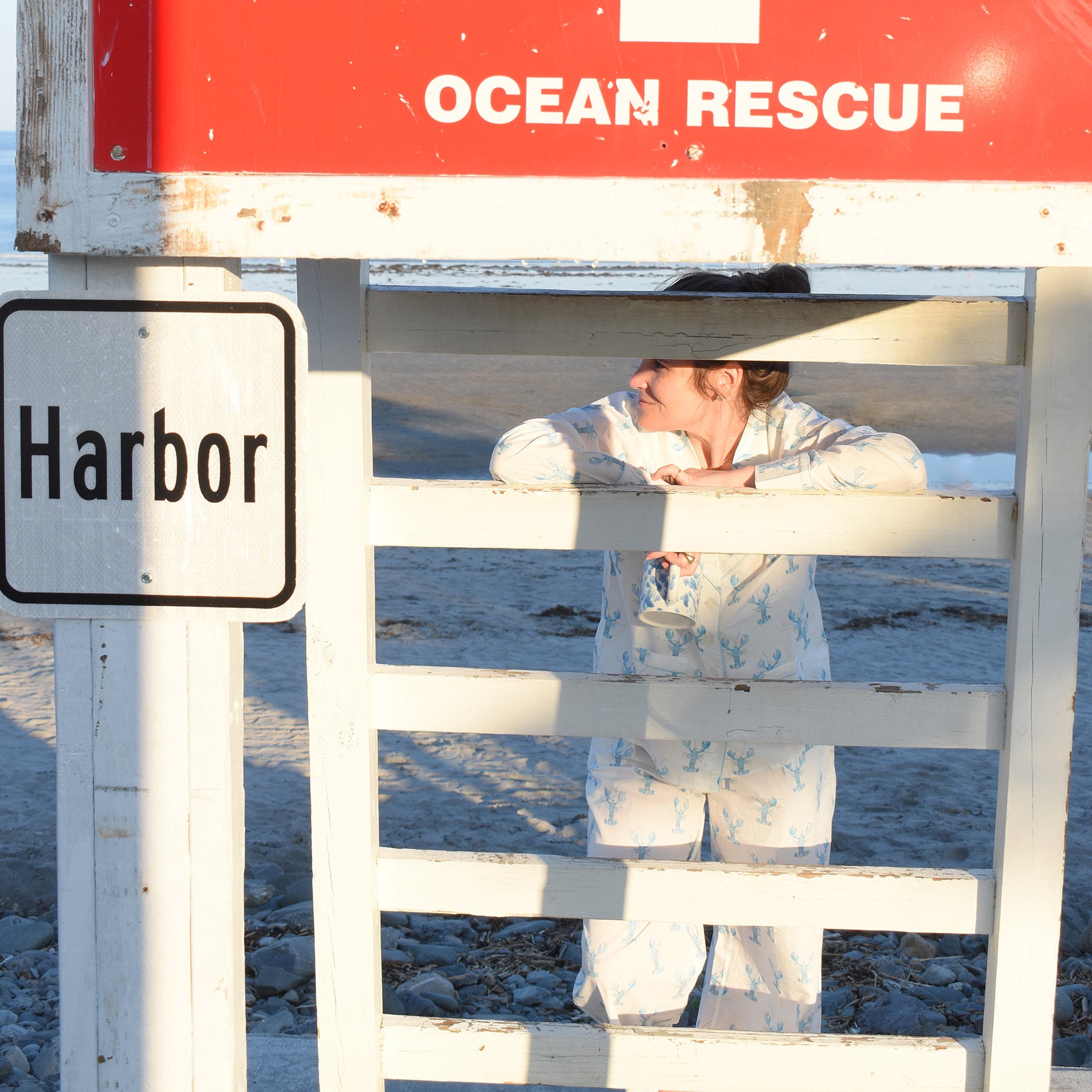 Wearing the Sara Fitz Blue Lobster Pants Pajama Set, a woman leans on the railing of a white lifeguard tower marked OCEAN RESCUE, near a Harbor sign, gazing to the side on a sandy beach.