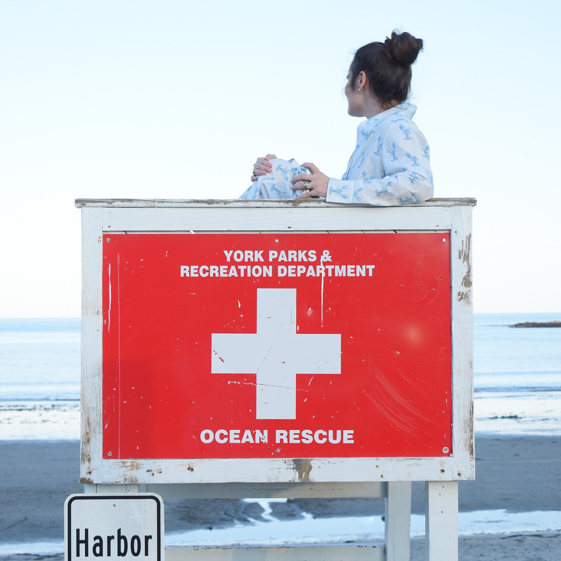 Wearing the Sara Fitz Blue Lobster Pants Pajama Set, a person sits on a lifeguard chair marked “York Parks & Recreation Department Ocean Rescue,” gazing out at the peaceful beach and ocean.