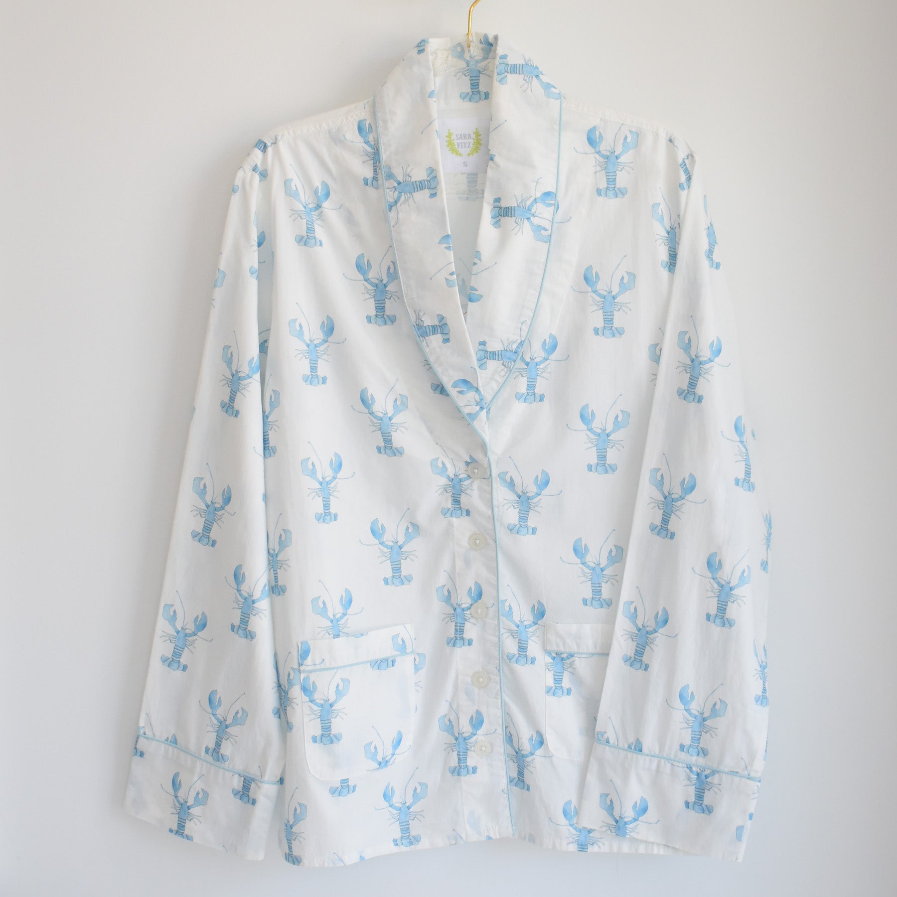 White pajama top with light blue lobster print, hanging on a gold hanger against a plain white background. Part of the Sara Fitz Blue Lobster Pants Pajama Set, this shirt features a collar, button front, and two front pockets—crafted from 100% cotton for breathable comfort and timeless coastal sleepwear style.