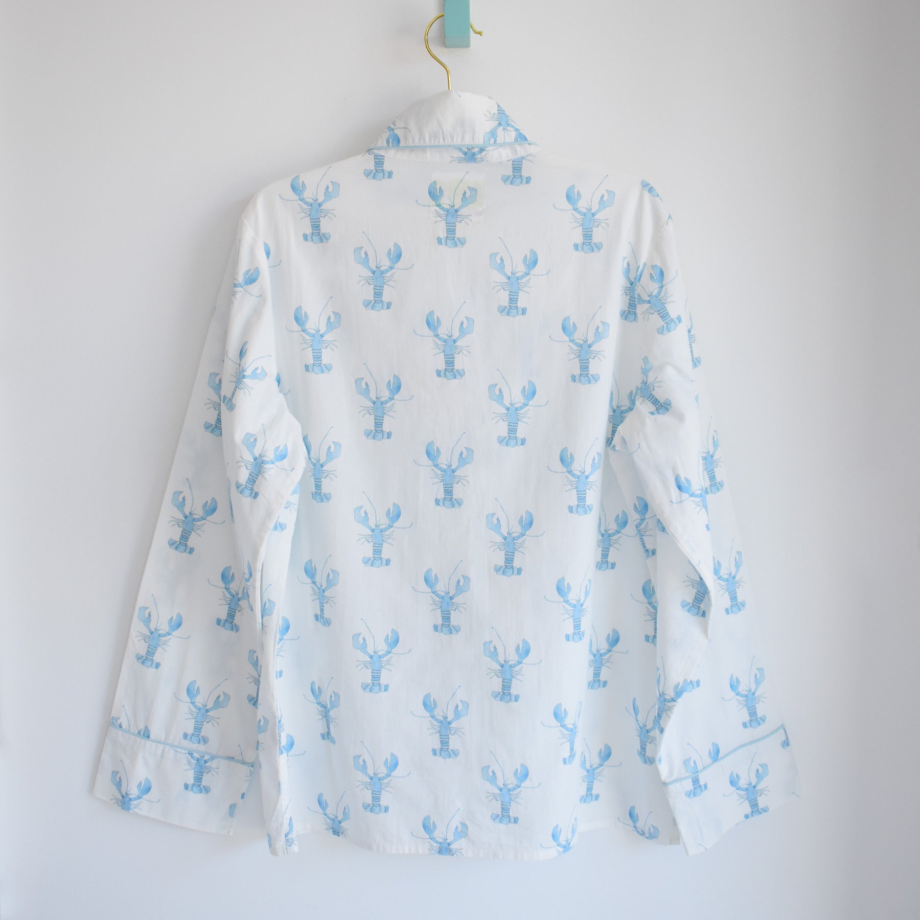 A white long-sleeve button-up shirt hangs on a hanger, adorned with a repeating light blue lobster pattern reminiscent of Sara Fitz style. Shown from the back against a plain white background, this piece pairs perfectly with our Sara Fitz Blue Lobster Pants Pajama Set for a stylish blend of comfort and seaside charm.