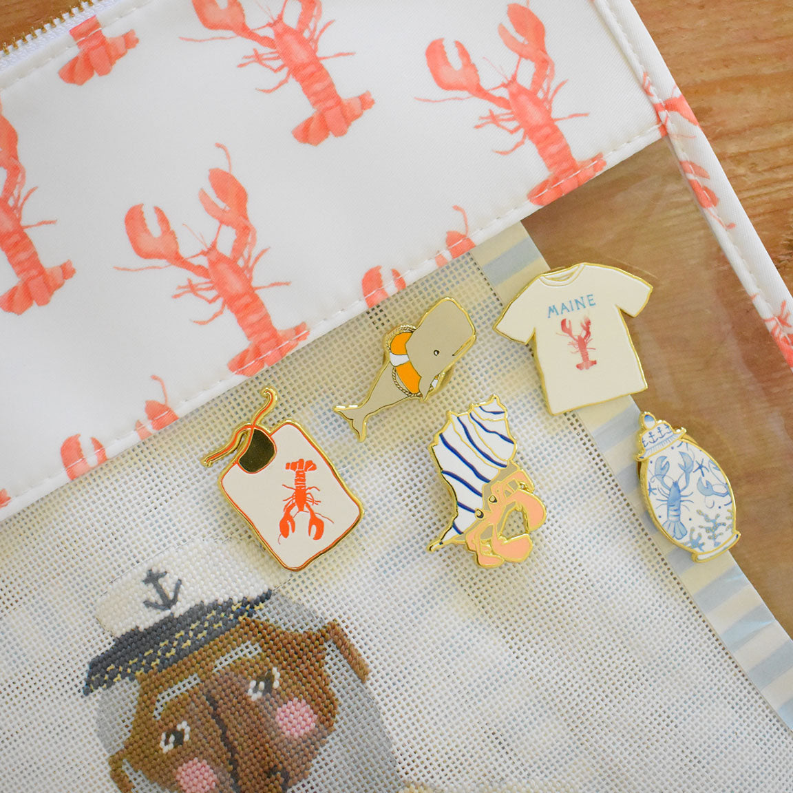 Five enamel pins from Sara Fitz—a lobster, whale, Maine t-shirt, doughnut hat, and blue seashell—are arranged on a mesh pouch. The Lobster Bib Needle Minder set is a perfect stitching accessory or enamel needle minder, bringing charming flair to your collection. The background features a playful lobster print and part of a dog illustration in a sailor hat.