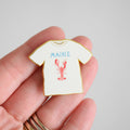A hand holds the Sara Fitz Maine Tee Needle Minder, a small enamel pin shaped like a white T-shirt featuring MAINE in blue letters and a red lobster graphic—perfect as an enamel needle minder or stitching accessory for your next project.
