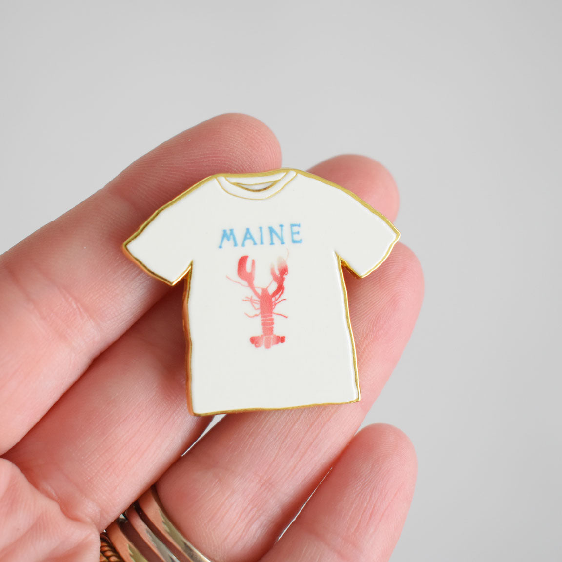 A hand holds the Sara Fitz Maine Tee Needle Minder, a small enamel pin shaped like a white T-shirt featuring MAINE in blue letters and a red lobster graphic—perfect as an enamel needle minder or stitching accessory for your next project.
