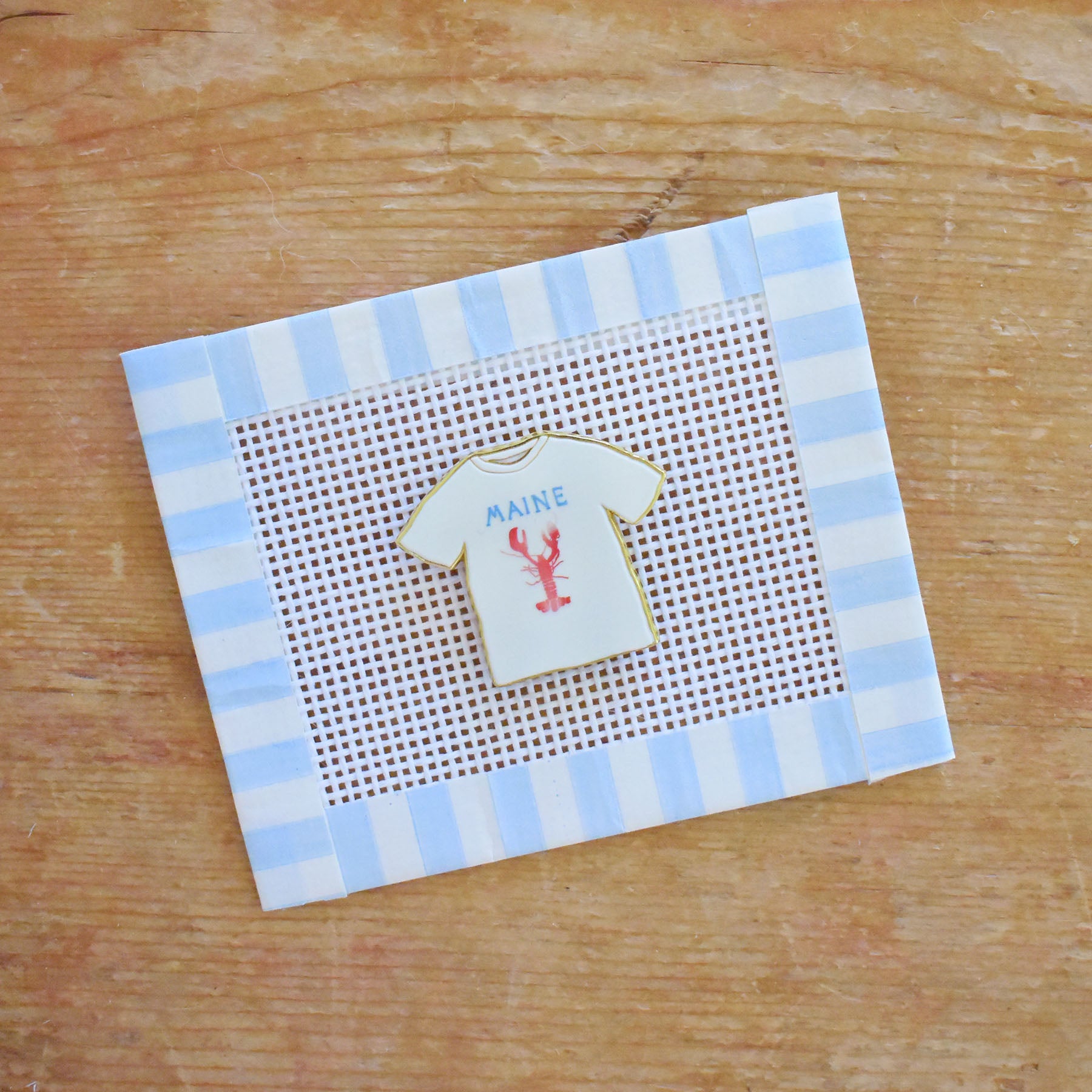 Introducing the Maine Tee Needle Minder by Sara Fitz—a charming enamel accessory shaped like a white T-shirt featuring “MAINE” and a red lobster. Placed on white mesh and bordered with a blue and white striped frame atop a wooden surface, this delightful piece is the perfect stitching companion for any needle holder.