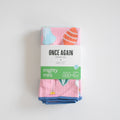 A folded pink Sara Fitz Mighty Mini Towel, Ornament, with a festive print wrapped in a white and green product sleeve.