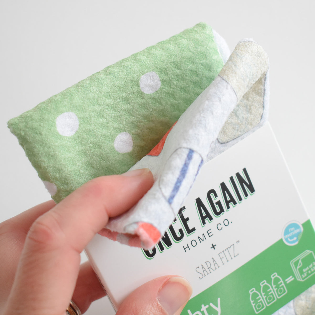A hand holds a folded green-and-white polka dot Mighty Mini Towel over its product packaging.