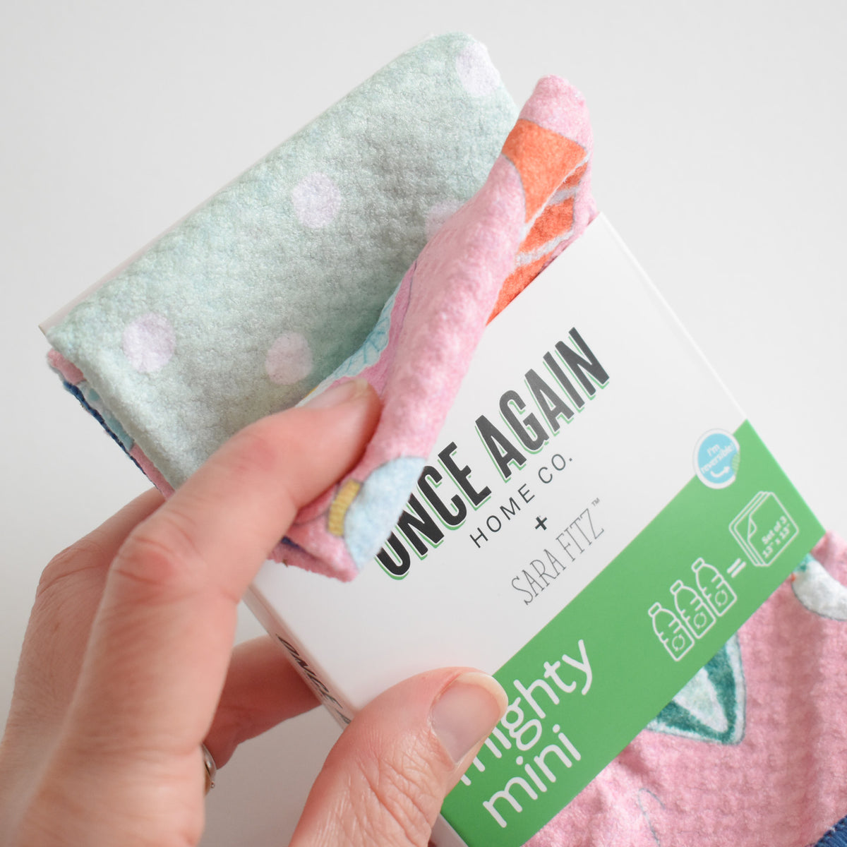 A hand holds a stack of three small, patterned Mighty Mini Towel, Ornament kitchen towels in a branded Sara Fitz cardboard sleeve.
