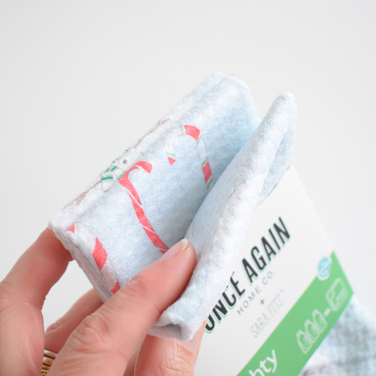 A hand holds a folded, light blue Mighty Mini Towel, Happy Snowman, featuring a candy cane pattern and secured with a green paper band.