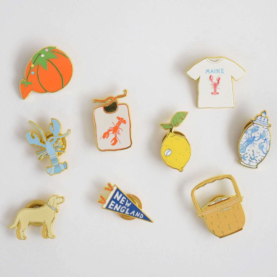 Showcased on a white background, the Blue Lobster Needle Minder by Sara Fitz is featured alongside nine whimsical enamel pins—a tomato, lobster, lobster on a luggage tag, lemon, blue can, yellow dog, “New England” pennant, picnic basket, and a Maine t-shirt with a red lobster. These playful designs make perfect stitching accessories or unique enamel needle minders for all your crafting projects.