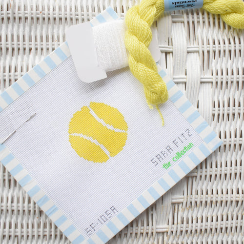 The MINI Tennis Ball Needlepoint by Sara Fitz features a yellow tennis ball on 18 mesh white canvas, with yellow and white threads above it, framed by blue and white stripes on a white wicker background.