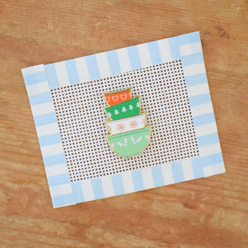 The Mixing Bowls Needle Minder by Sara Fitz features an illustration of four vintage-patterned orange and green bowls, stacked on a brown wooden surface, all framed by a blue and white striped border.