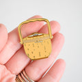 A hand holds the Sara Fitz Nantucket Basket Needle Minder, a small enamel pin shaped like a yellow picnic basket with a handle and clasp, perfect as both an adorable stitching accessory and needle minder, against a plain light background.