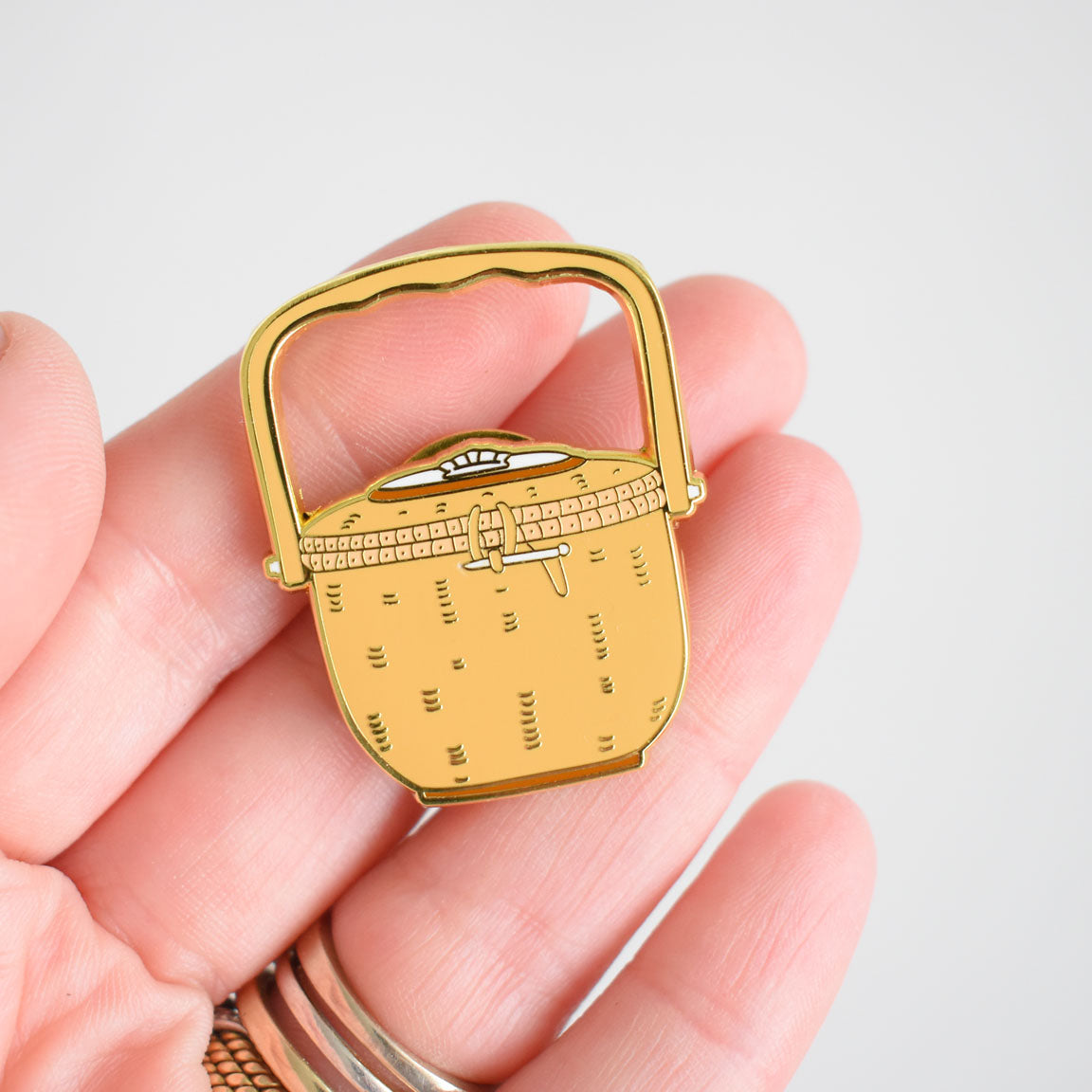 A hand holds the Sara Fitz Nantucket Basket Needle Minder, a small enamel pin shaped like a yellow picnic basket with a handle and clasp, perfect as both an adorable stitching accessory and needle minder, against a plain light background.