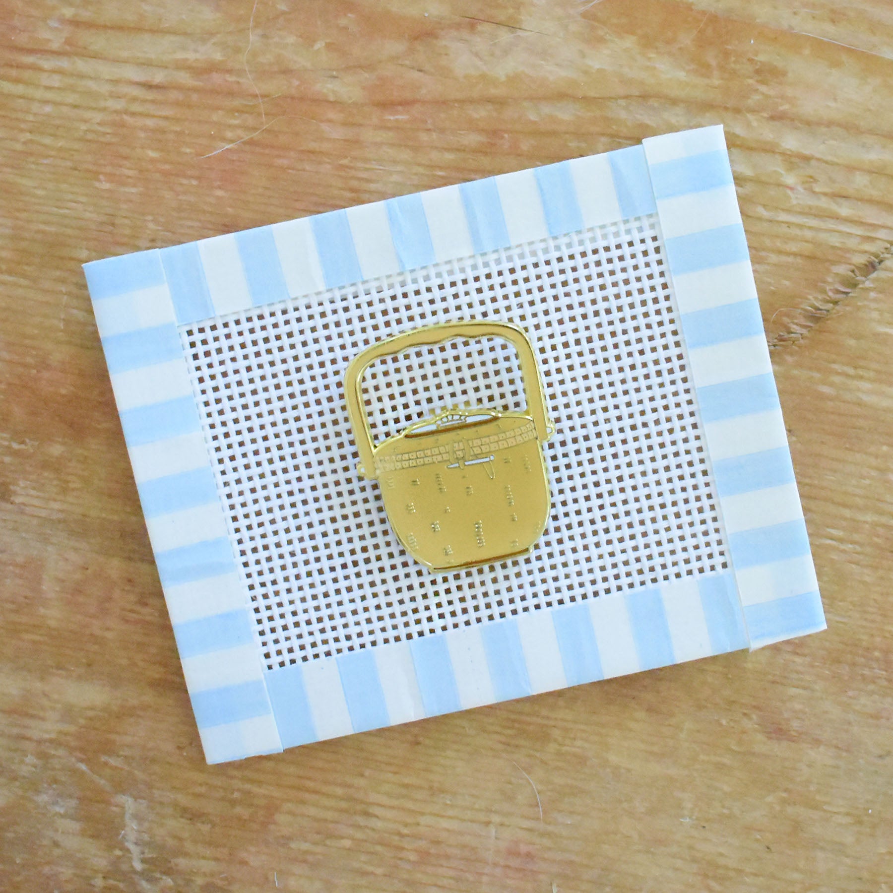 The Sara Fitz Nantucket Basket Needle Minder—a gold bag-shaped pin designed as a stitching accessory or needle holder—rests on a square piece of mesh fabric bordered with blue and white striped tape, displayed atop a wooden surface.