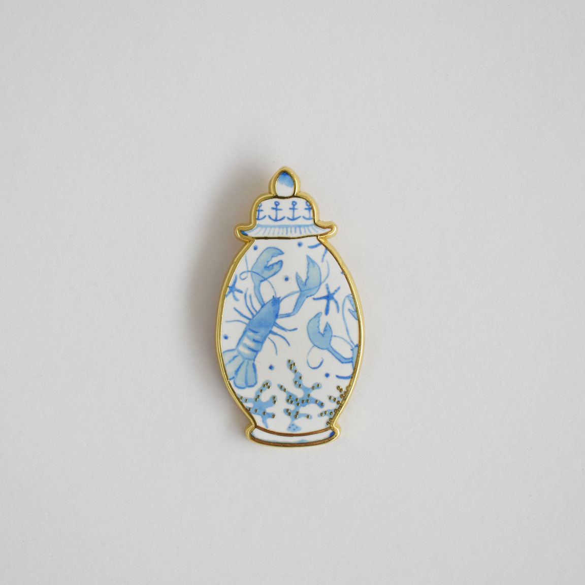 Introducing the Ginger Jar Needle Minder by Sara Fitz—a decorative enamel pin inspired by a classic vase shape. It features delicate blue line art of a lobster and seaweed on a crisp white background, all outlined in gold. Designed to keep your needles secure and stylish while you stitch, this unique piece is both an elegant accessory and practical tool for any crafter.