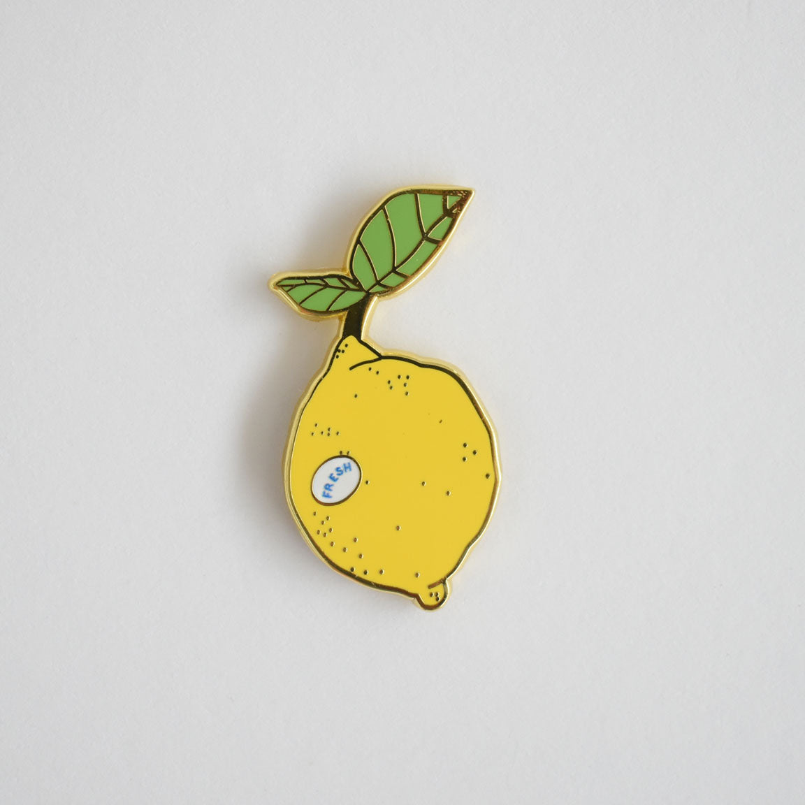 The Lemon Needle Minder by Sara Fitz is a yellow enamel pin shaped like a lemon, featuring a green leaf and accented with a small blue sticker that reads FRESH. Displayed on a plain white background, this charming accessory doubles as an enamel needle minder—perfect for keeping your needles secure and within reach while you stitch.