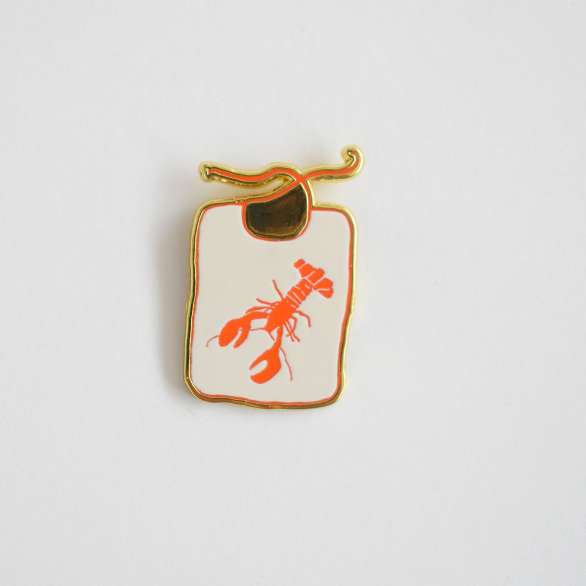 The Sara Fitz Lobster Bib Needle Minder is a charming enamel accessory shaped like a white cutting board with a gold outline, featuring an orange lobster illustration in the center and a gold handle at the top. This stylish needle minder keeps your needles secure while you stitch.