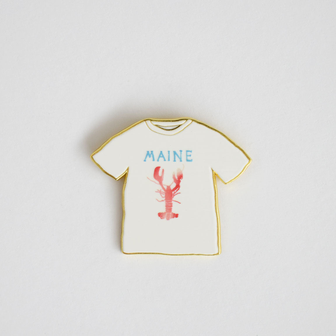 The Maine Tee Needle Minder by Sara Fitz is a white T-shirt-shaped stitching accessory featuring "MAINE" in blue text and a red lobster illustration outlined in gold, displayed against a plain white background.
