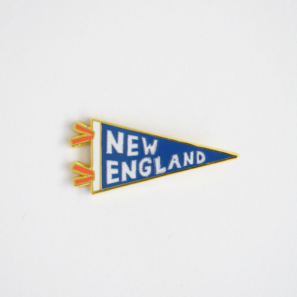 A blue Pennant Needle Minder by Sara Fitz, featuring yellow edging, red ties, and bold white "NEW ENGLAND" text on its surface, set against a simple white background—a stylish enamel accessory to keep your needle holder secure or add a charming touch to any stitching enthusiast’s collection.