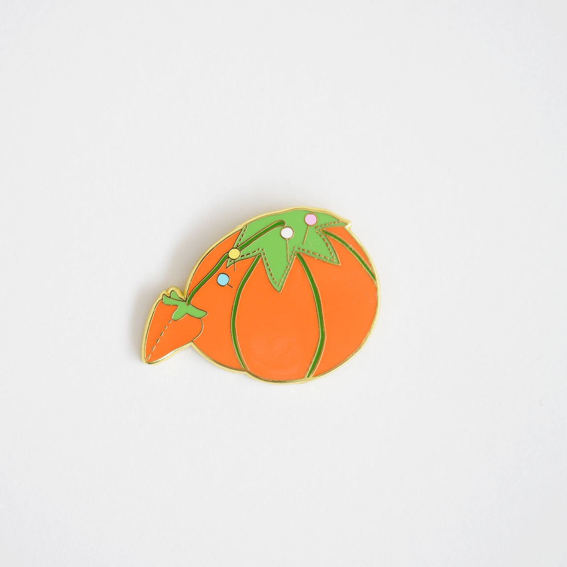 The Sara Fitz Tomato Pincushion Needle Minder, featuring an enamel pin shaped like an orange tomato with green leaves and tiny white dots, is displayed on a plain white background and makes a charming stitching accessory for any crafter’s collection.