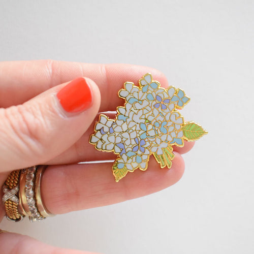 A hand with orange nail polish and rings holds the Sara Fitz Hydrangea Bloom Needle Minder—an enamel pin shaped like light blue and white hydrangea flowers with green leaves, perfect as a charming stitching accessory for your craft projects.
