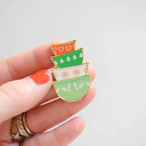 A hand with orange nail polish holds the Sara Fitz Mixing Bowls Needle Minder, a festive accessory shaped like four stacked bowls adorned in red, green, and white holiday patterns—perfect for cheerful stitching projects.