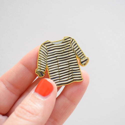 A hand with orange nail polish holds the Sara Fitz Striped Shirt Needle Minder, a small enamel accessory shaped like a long-sleeved black-and-white striped shirt, against a plain light background.
