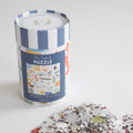 A cylindrical canister containing a 500-piece New England Puzzle, with a pile of loose pieces from the nostalgic collage in the foreground.