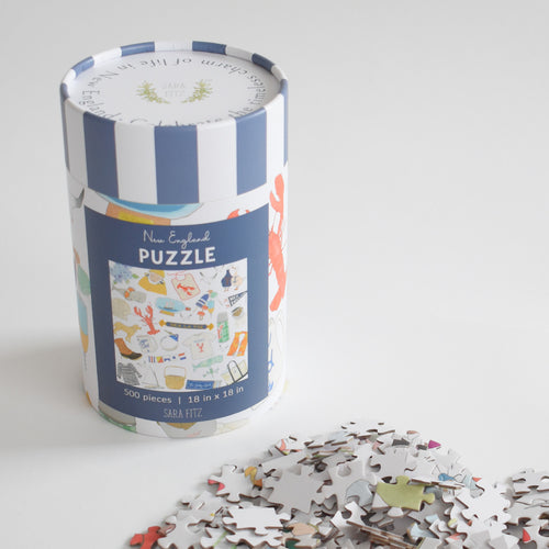 A cylindrical canister containing a 500-piece New England Puzzle, with a pile of loose pieces from the nostalgic collage in the foreground.