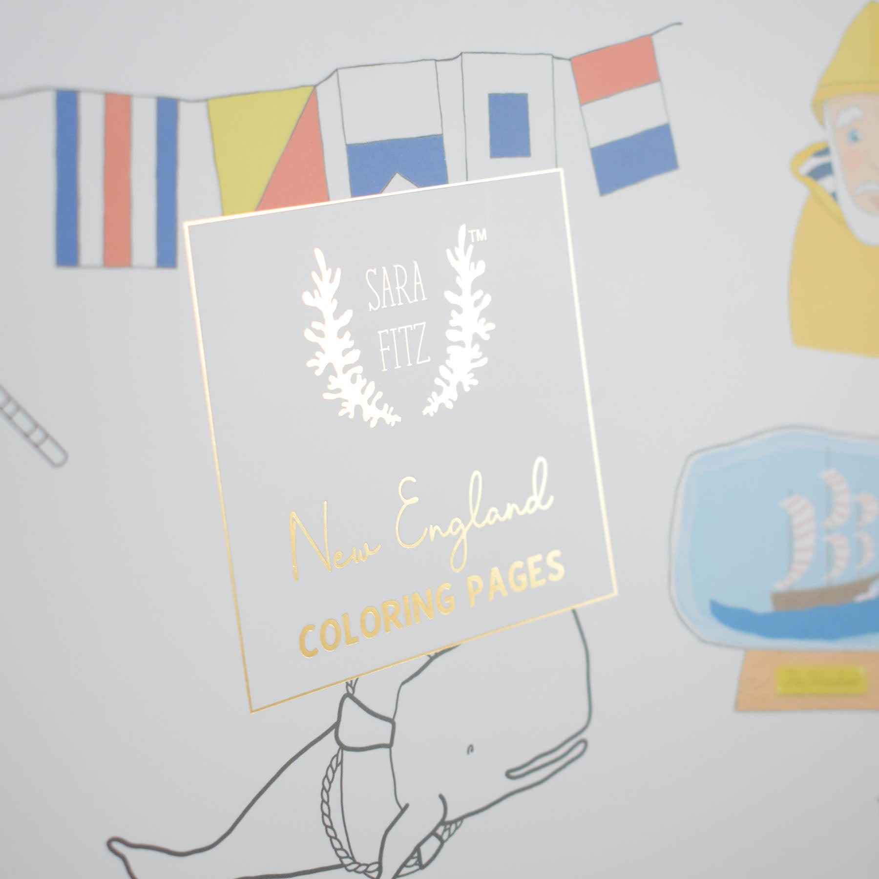 A sticker in gold and white with “Sara Fitz Coloring Book New England” covers part of a nautical-themed coloring page featuring classic New England illustrations—such as flags, a whale, a ship in a bottle, and a yellow figure—making it the perfect addition to any Sara Fitz coloring book or adult coloring collection.