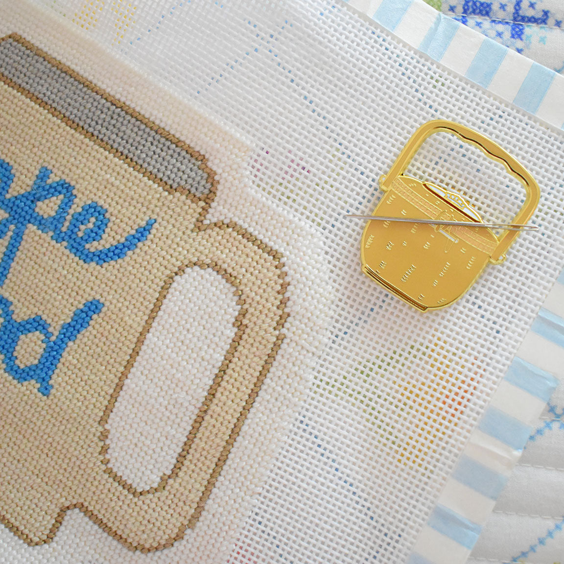A cross-stitch project featuring part of a beige mug with blue text is displayed on white mesh fabric trimmed with a striped border. Beside it lies the Nantucket Basket Needle Minder by Sara Fitz—a gold enamel accessory shaped like a kettle and holding a needle—making it the perfect stitching companion.