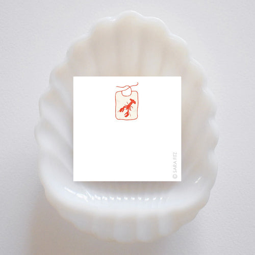 A white, scallop-shaped dish holds a square notelet featuring a simple red line drawing of a lobster bib on its smooth white surface. The artist’s signature, Sara Fitz, appears in the lower right corner. Printed in the USA.