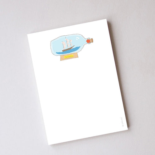 The Sara Fitz Boat in a Bottle Notepad is 5x7 inches with an illustrated ship in a glass bottle on a stand near the top, leaving space for notes. Made in the USA.