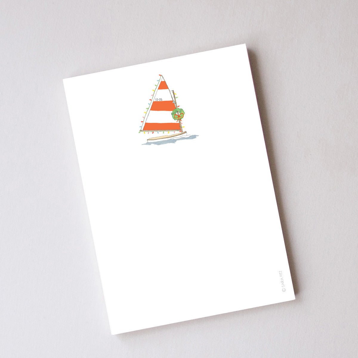 A Holiday Sailboat Notepad featuring a sailboat with an orange and white striped sail decorated with Christmas lights, a wreath, and the date 12-25.