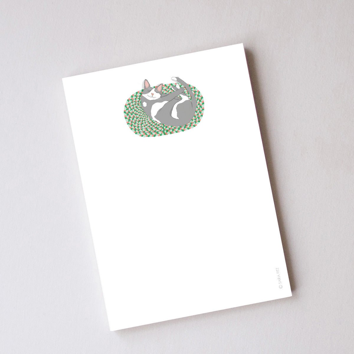 A Cozy Kitty Notepad featuring an illustrated grey and white cat curled up on a patterned rug, containing unlined pages.