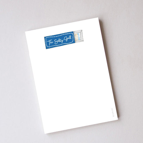 The Sara Fitz Matchbox Notepad, made in the USA, features a white cover illustrated with a blue "The Salty Grill" matchbox at the top and offers blank, unlined pages for your notes.