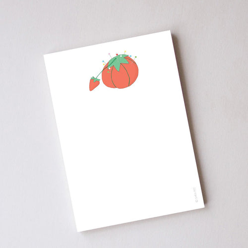 The Sara Fitz Tomato Pincushion Notepad, made in the USA, displays a red tomato pincushion and strawberry illustration at the top, with blank tear-off pages ideal for notes or lists.