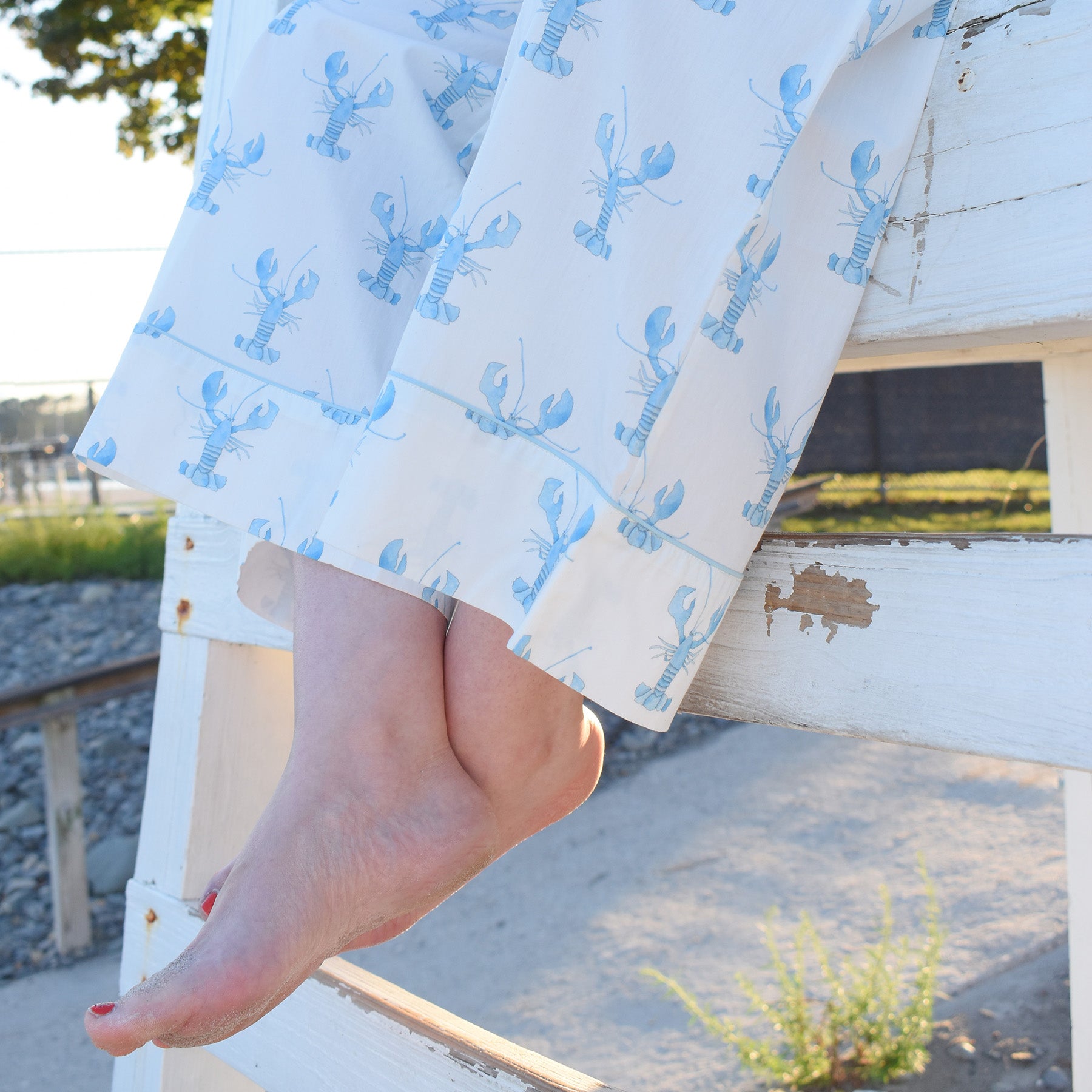 Bare feet with red nail polish rest on a white wooden structure. The person is wearing the Blue Lobster Pants Pajama Set from Sara Fitz, crafted from 100% cotton—perfect coastal sleepwear. Sunlight casts gentle shadows while rocks and greenery are visible in the background.